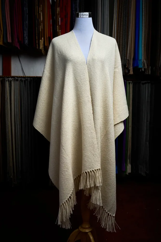 Ruana de baby alpaca color crudo con flecos, sobre maniquí y fondo oscuro