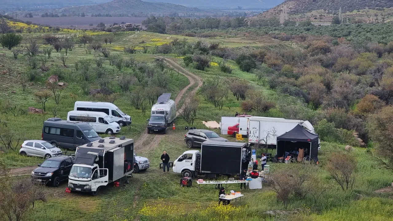 Vista aérea del campamento base de un rodaje en exterior: furgones y camiones de equipo estacionados junto a un camino de tierra, una carpa negra, mesas de alimentación y personas del equipo entre los vehículos, en un valle con cerros al fondo